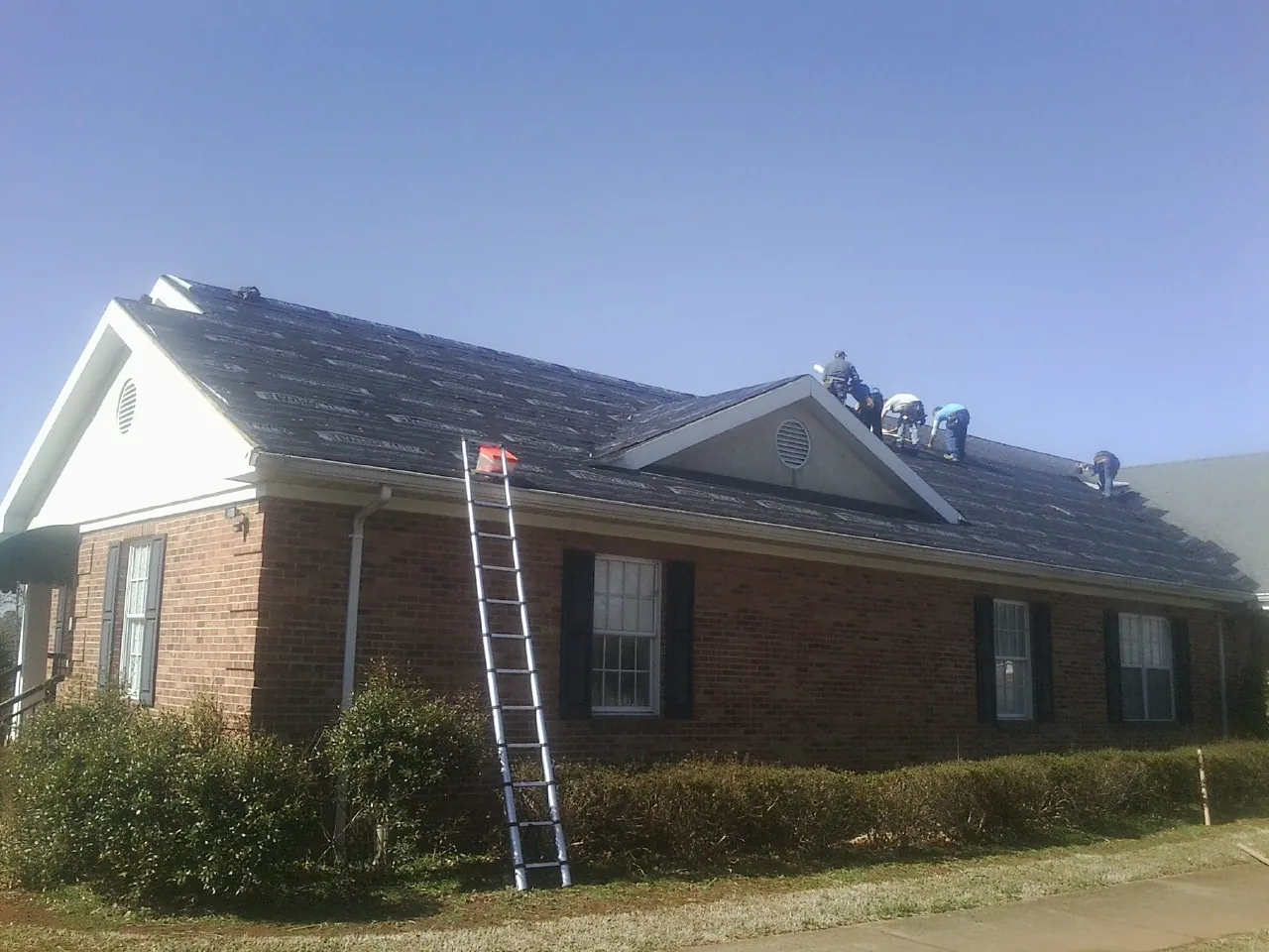Crew tearing shingles off a brick ranch roof with a ladder set — Metal Roof Repair work by our crew in Loudon