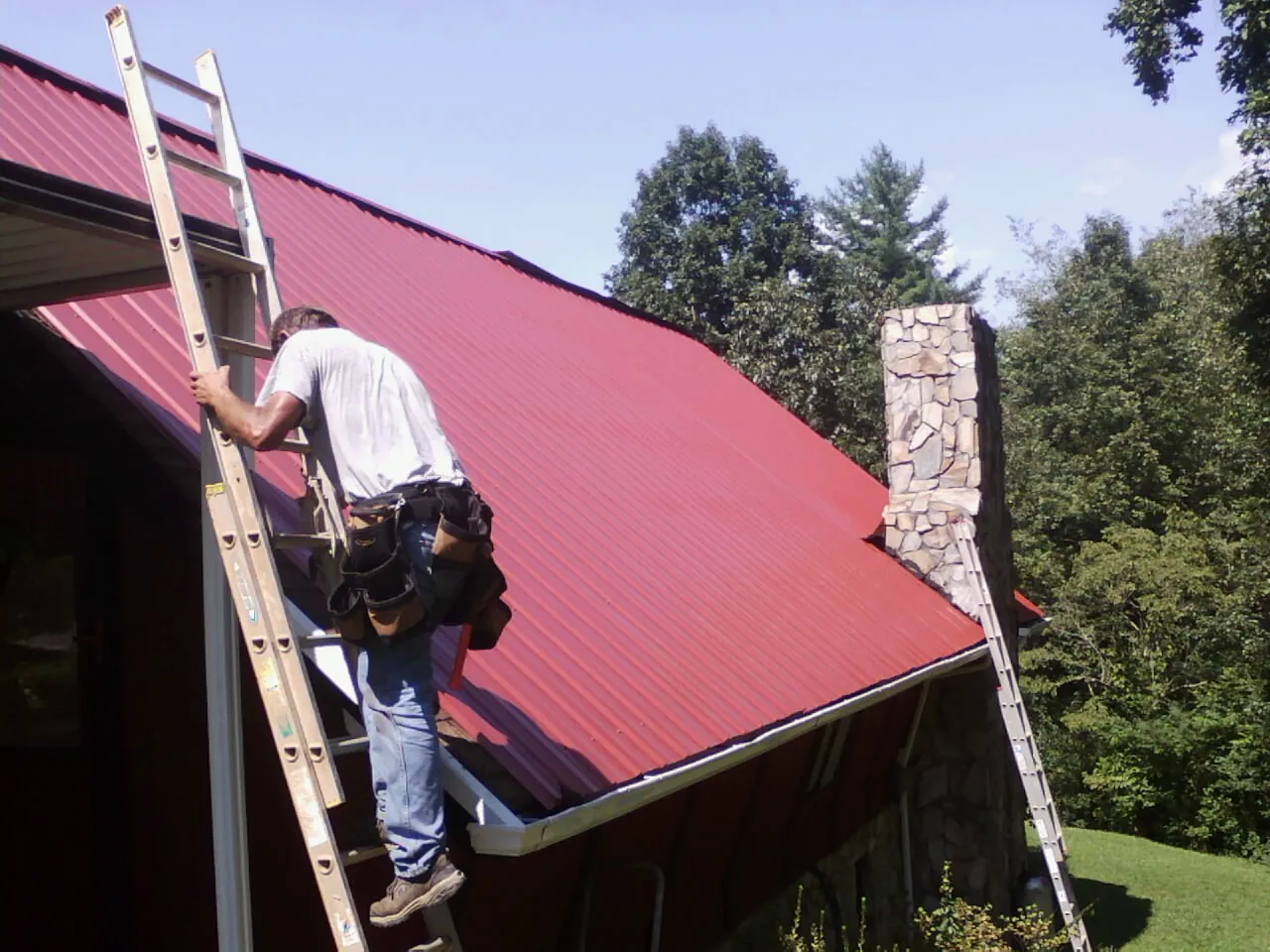 Roofer on a ladder beside a red metal roof and stone chimney — Soffit & Fascia Repair by Roofing Brothers in Loudon